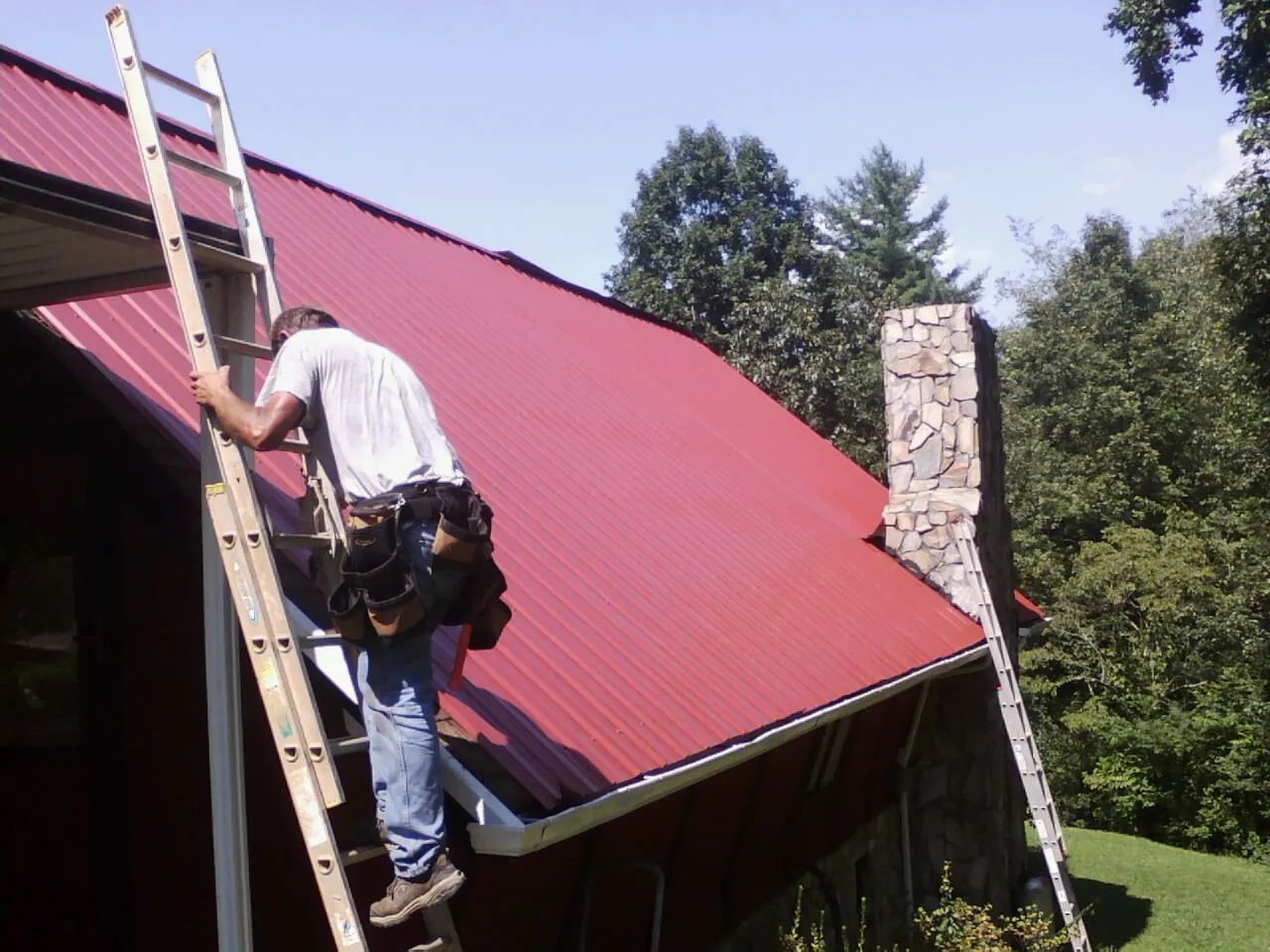 Roofer on a ladder beside a red metal roof and stone chimney — Soffit & Fascia Repair by Roofing Brothers in Oklahoma City